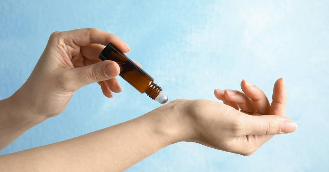 A woman’s arms against a blue backdrop. She uses one hand to apply an essential oil roller onto the wrist of her other arm.