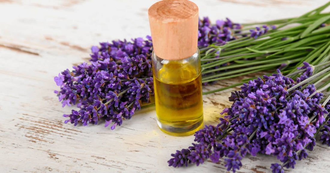 A glass vial with a wooden topper, filled with essential oils. The vial sits on a wood table beside lavender flowers.