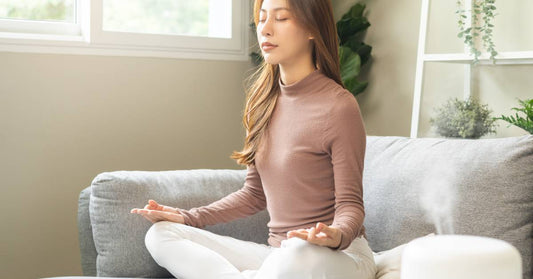 A young woman sitting on her couch, practicing meditation, while a nearby diffuser sprays essential oils in the air.